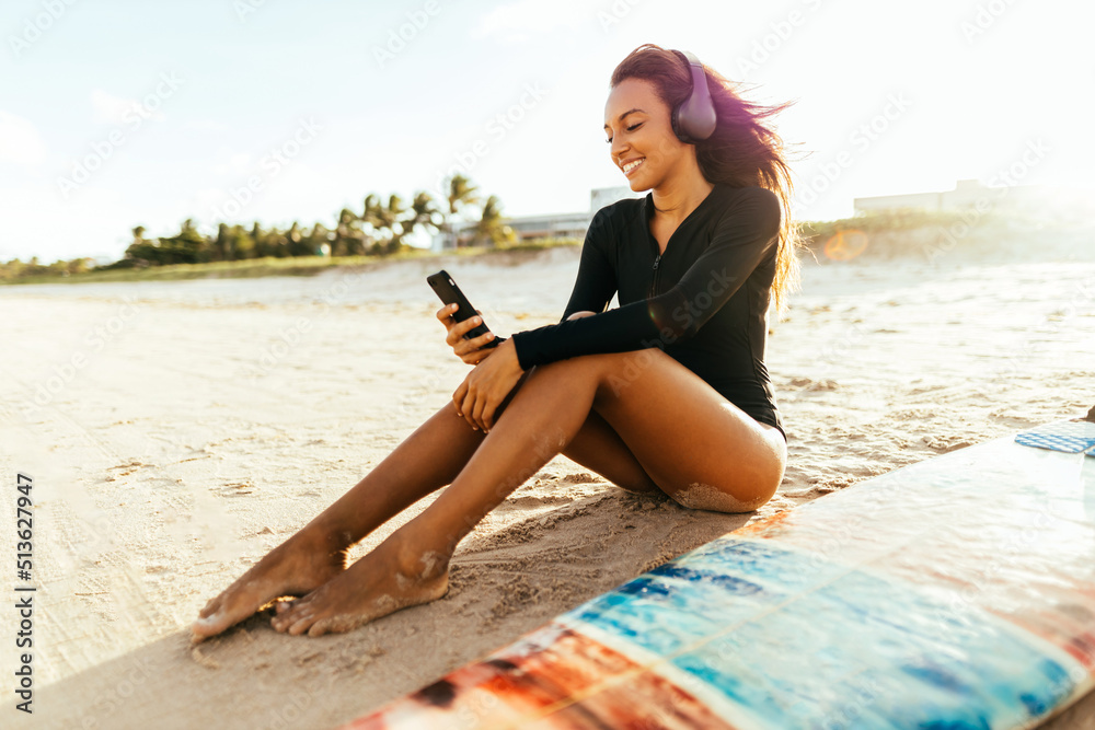 Portrait of young female surfer on the beach with her surfboard listening to music