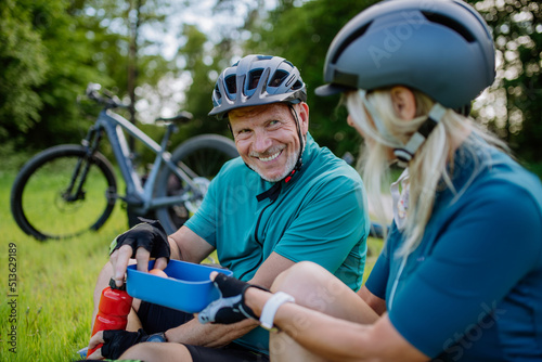 Active senior couple resting after bicycle ride at summer park, sitting on grass and having snack.