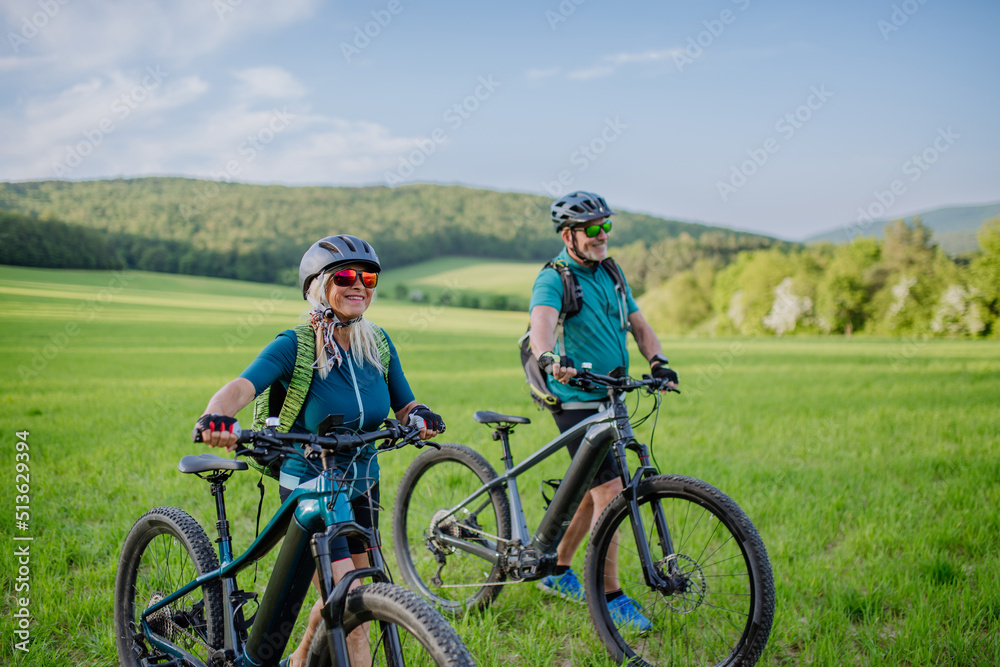 Active senior couple with electric bicycles on meadow in summer, healthy lifestyle concept.