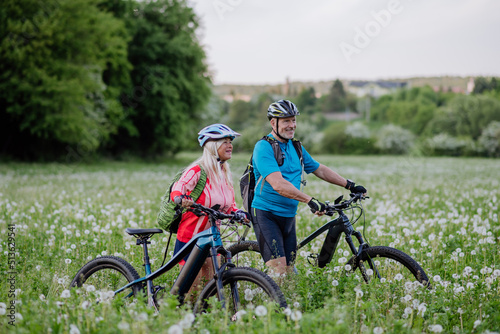 Active senior couple with electric bicycles on meadow in summer, healthy lifestyle concept.