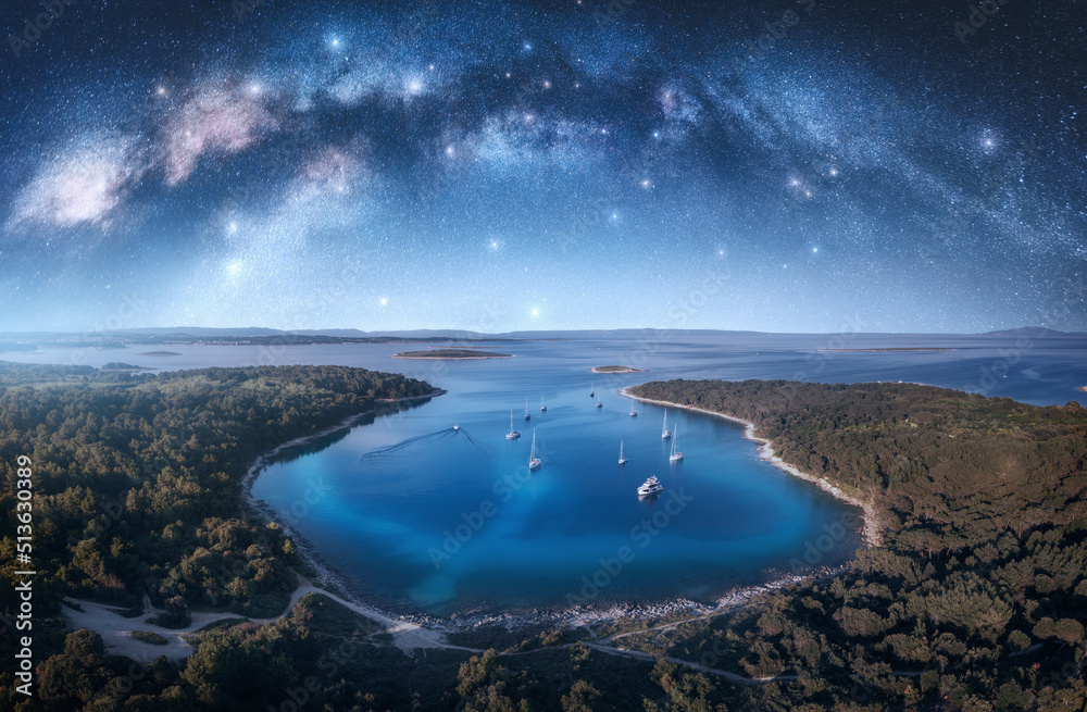 Milky Way arch and beautiful yachts and boats on the sea bay at summer night. Kamenjak, Croatia. Space. Top view of arched milky way, starry sky, sailboats, lagoon, clear blue water, forest. Travel