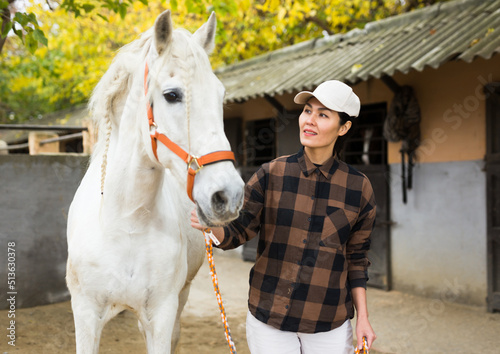 Asian woman horse breeder leading white horse through paddock.