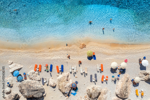 Fototapeta Naklejka Na Ścianę i Meble -  Aerial view of blue sea, sandy beach with umbrellas, stones and rocks, clear water at sunset in summer. Lefkada island, Greece. Tropical landscape with sea shore, swimming people, blue water. Top view