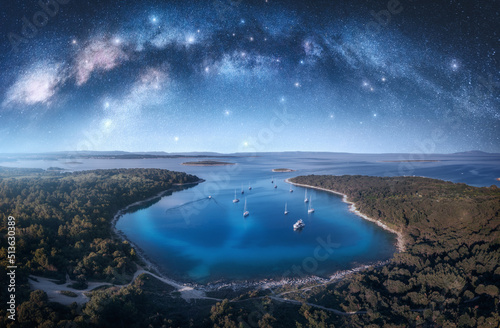 Milky Way arch and beautiful yachts and boats on the sea bay at summer night. Kamenjak, Croatia. Space. Top view of arched milky way, starry sky, sailboats, lagoon, clear blue water, forest. Travel © den-belitsky