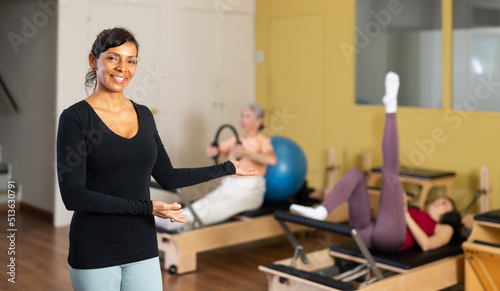 Portrait of friendly Hispanic female fitness instructor smiling at camera and inviting to pilates reformer studio