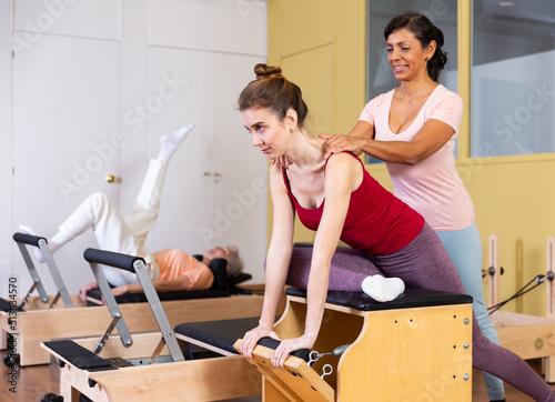 Fototapeta Young European woman practicing Pilates in a group workout performs an exercise on a combined chair, where a latin american
