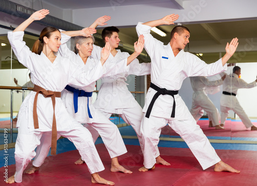 Various aged men and women in white kimono and belts training kata moves in gym.