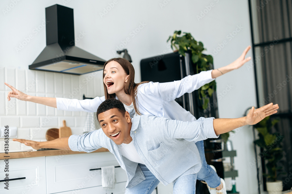 Fototapeta premium Multiracial happy couple in love, caucasian girl and latino guy, dressed in a modern casual clothes, have fun together at home in the kitchen, laugh, enjoy the weekend, experiencing positive emotions