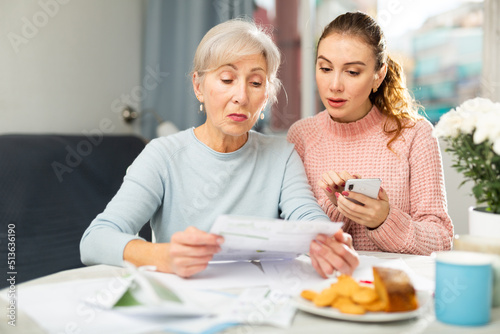 Interested focused elderly woman sitting in dining room with adult daughter using smartphone and checking utility bills.