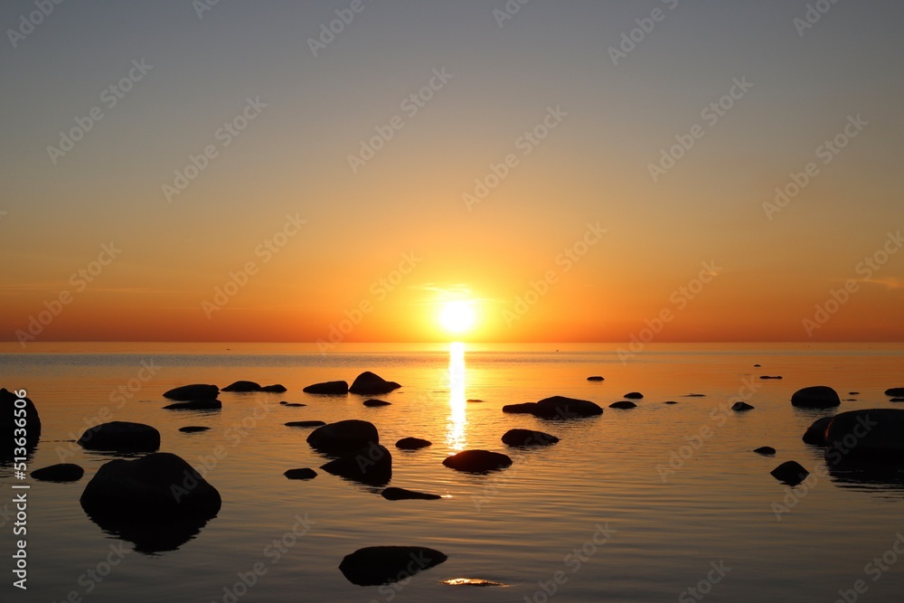 Sunset over calm Baltic sea, in Saulkrasti, Latvia. Sun shining from behind clouds, bright reflection on water, black shapes of boulders. Selective focus