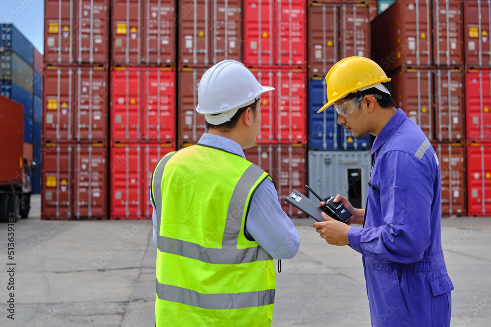 Two professional Asian male workers in safety uniforms and hard hats work at a logistics terminal with many stacks of containers, loading control shipping goods for the cargo transportation industry. Two professional Asian male workers in safety uniforms and hard hats work at a logistics terminal with many stacks of containers, loading control shipping goods for the cargo transportation industry.