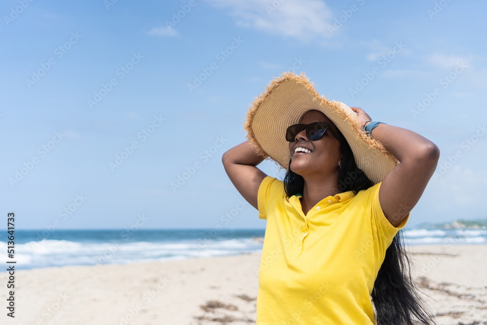 Cheerful young female in summer hat and sunglasses touching head and looking away with smile while relaxing against sea on sunny weekend day