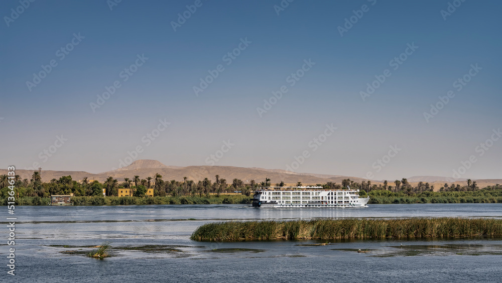 There are islands with green vegetation in the riverbed. A white cruise ship is sailing on blue water. A house is visible among the palm trees on the shore. Sand dunes against the sky. Egypt. Nile