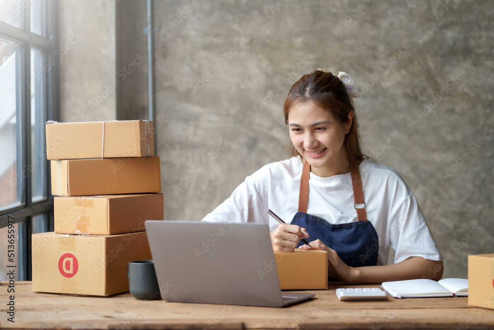 Happy young Asian business owner woman prepare parcel box and check online orders of product for deliver to customer on laptop computer. Shopping Online concept. Happy young Asian business owner woman prepare parcel box and check online orders of product for deliver to customer on laptop computer. Shopping Online concept.