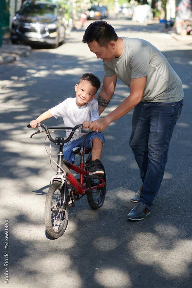 Happy father teaching smiling little son how to ride bicycle on sunny summer day Happy father teaching smiling little son how to ride bicycle on sunny summer day