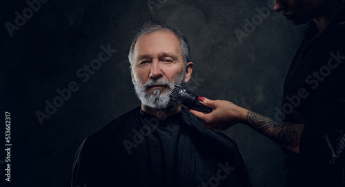 Shot of female barber with tattooed hand cutting elderly man using clipper against dark background. © Fxquadro