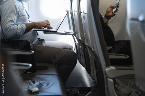 Businessman using laptop in airplane and passenger in front row with cellphone. Airline wifi connection, device flight mode concepts