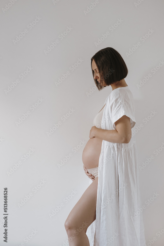 Beautiful happy pregnant woman in elegant white robe touching her belly tummy. Aesthetic elegant pregnancy, maternity, newborn baby expecting concept Beautiful happy pregnant woman in elegant white robe touching her belly tummy. Aesthetic elegant pregnancy, maternity, newborn baby expecting concept