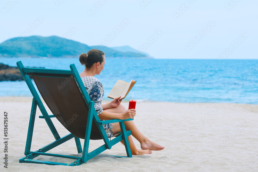 Woman sitting in deck chair at beach reading book on sunny day.