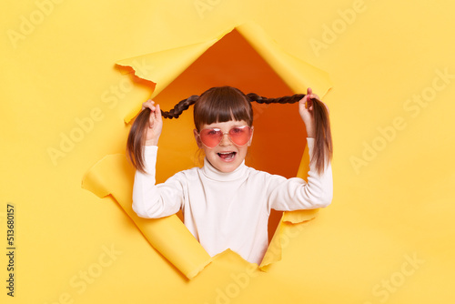 Portrait of excited amazed little girl with braids wearing white turtleneck and heart shape sunglasses posing in torn hole of yellow paper wall, standing raised her pigtails, keeps mouth opened.