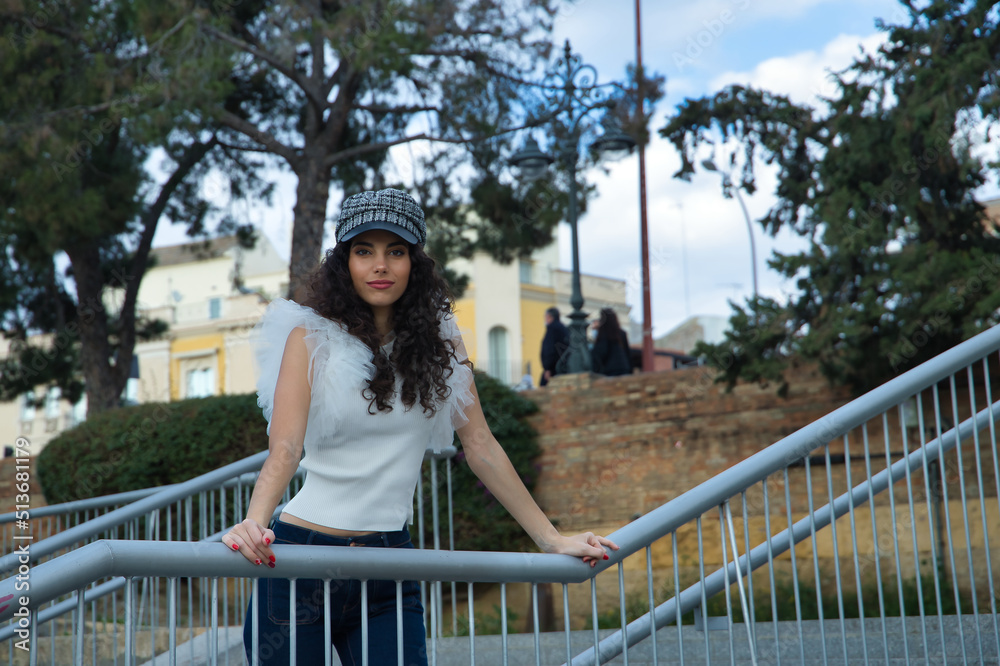 Young and beautiful woman, brunette, with curly hair, wearing white shirt, jeans and cap, leaning on a railing. Concept beauty, fashion, model, trend, tranquility.