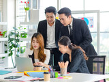 Asian millennial professional successful male businessman mentor in black formal suit standing helping two female employee sitting working with laptop computer at workstation in company meeting room