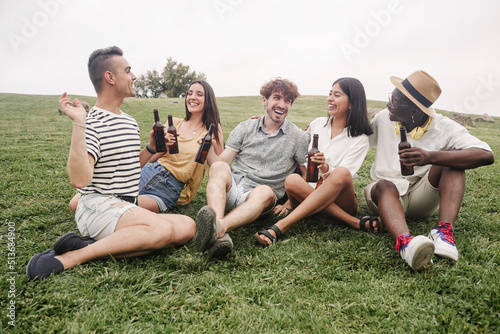 Group of young friends sitting on the grass having good times drinking beer in summer