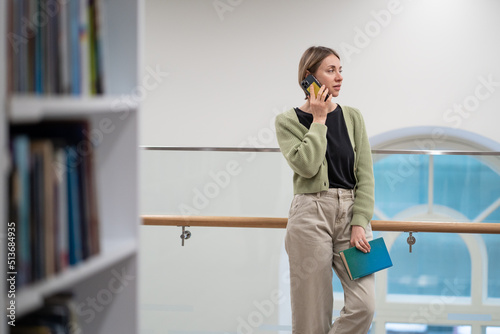 Female mature student talking on mobile phone in modern public library, middle-aged woman being distracted by phone call while choosing a book in bookstore. Education, self-education