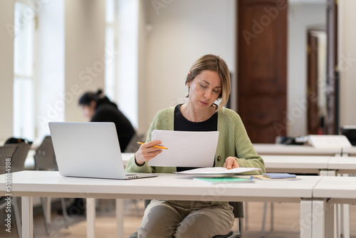 Tableau sur toile Focused hard-working middle-aged female student reading architectural drawings, working on graduation project while studying in public library