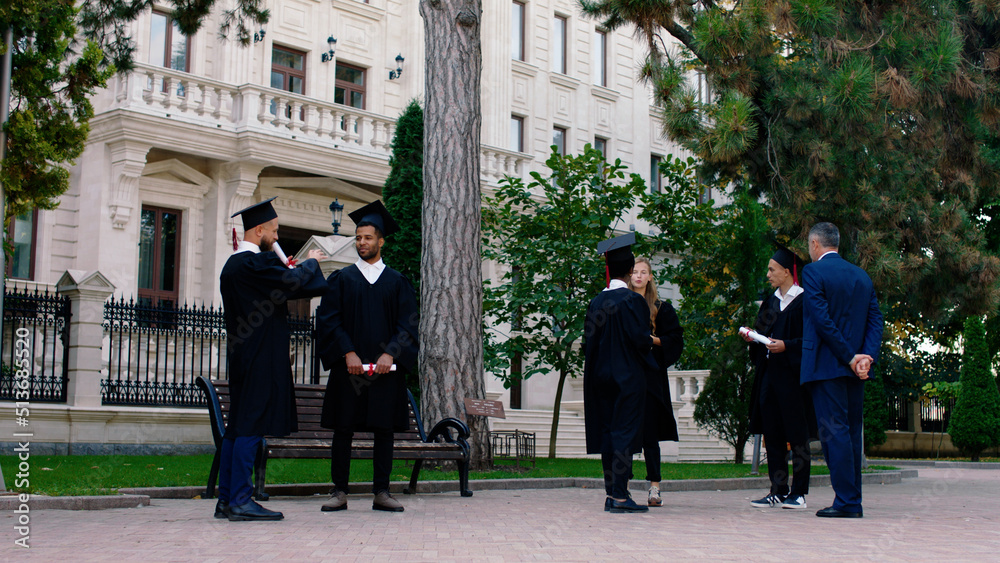 Obraz premium Multiethnic students after graduation meeting in the middle of college park and discussing they wearing graduation suits and caps