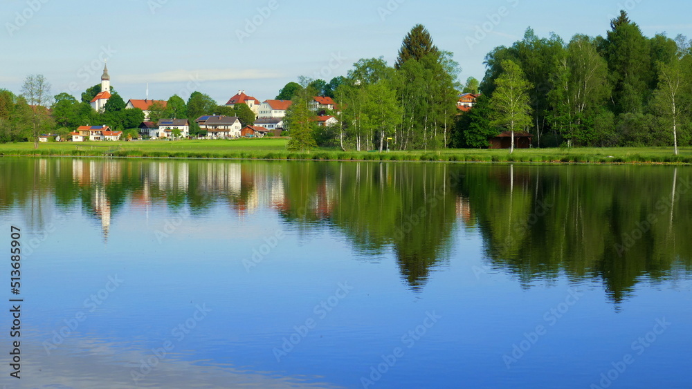 Blick über stillen Bad Soier See auf Bayersoien mit Spiegelung von Dorf und Wald unter blauem Himmel