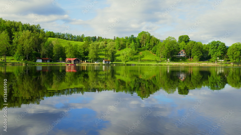 Blick über spiegelnden Bad Soier See mit grüner Landschaft und weißen Wolken bei blauem Himmel