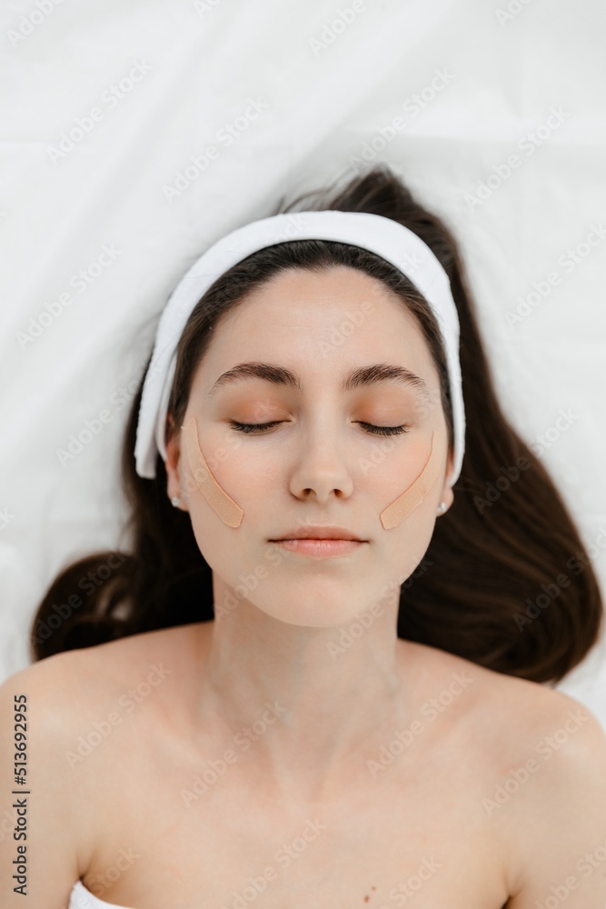 Cosmetology. beautiful young caucasian girl in the cosmetologist's office against a white background. A model with a headband and  with lifting tape on her face. Kinesio taping.