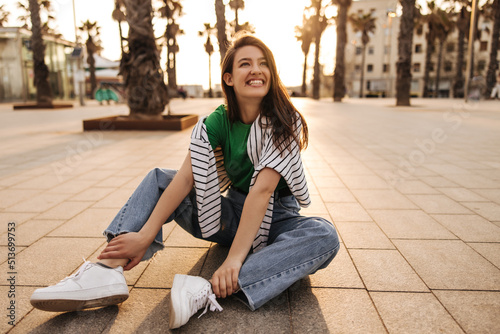 Cheerful young caucasian woman smiles with her teeth looking to side, sitting in European city on pavement. Brunette wears T-shirt, jacket and jeans. City life concept