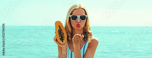 Summer portrait of young woman with papaya blowing her lips sending kiss on the beach on sea background