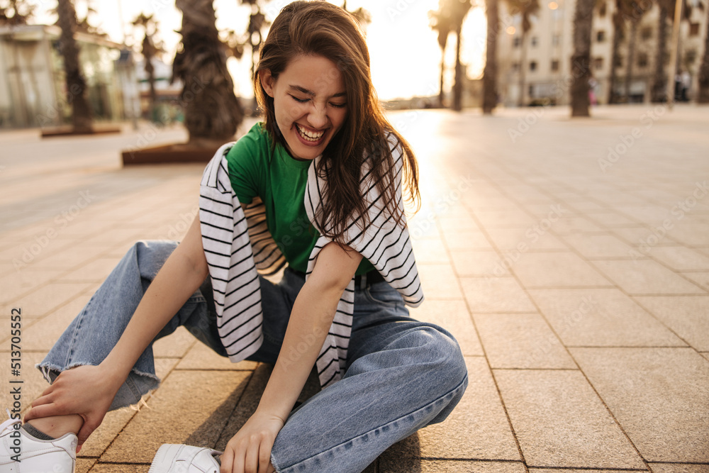 Funny young caucasian girl with closed eyes has fun sitting on pavement seeing off sunset. Brunette wears casual clothes in summer. Positive emotions concept