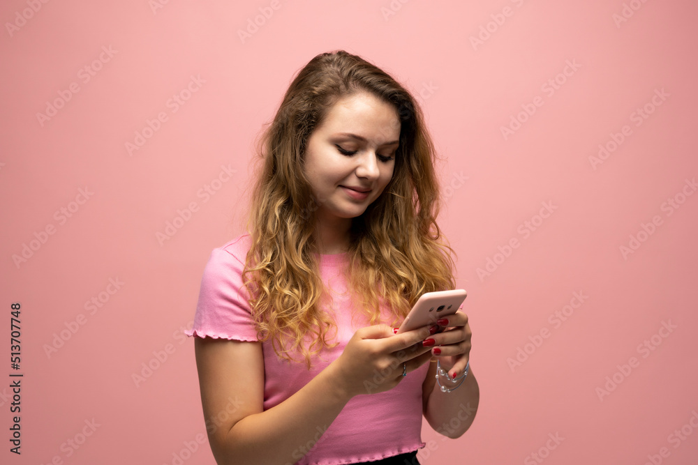 Smiling young woman girl in pink t-shirt using mobile phone and typing sms message to a friends isolated on pink wall background studio. People lifestyle concept. Mock up copy space. Smiling young woman girl in pink t-shirt using mobile phone and typing sms message to a friends isolated on pink wall background studio. People lifestyle concept. Mock up copy space.