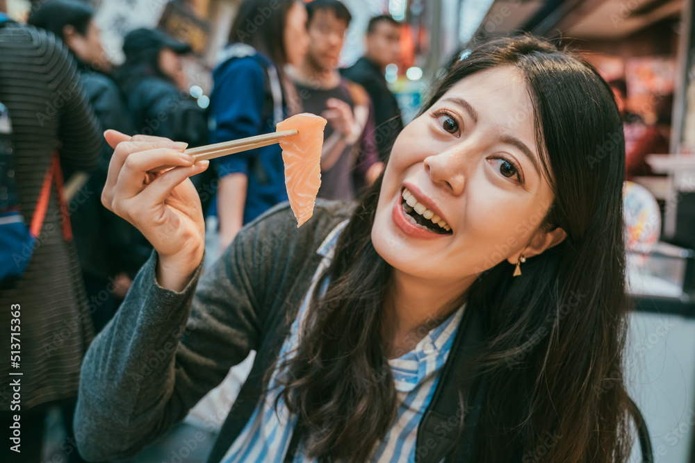 closeup portrait of pretty asian girl holding a piece of sashimi using chopsticks and looking at camera with smile on face in kuromon ichiba market in Osaka japan