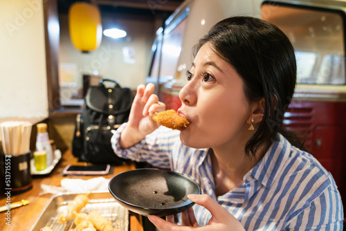 closeup of Japanese, Chinese, Korean girl holding small tray while eating biting in crispy tempura Japanese fried dish in an izakaya restaurant at dinner time in shinsekai new world Osaka japan