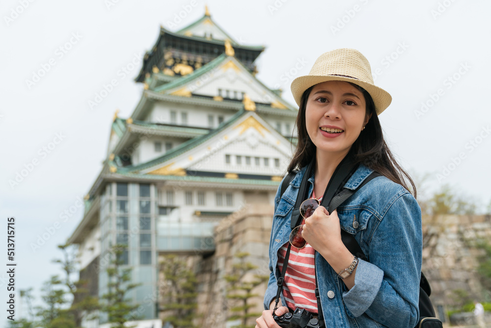 portrait of cheerful pretty Chinese girl backpacker wearing hat looking at camera against blurred background of Osaka castle in japan at springtime