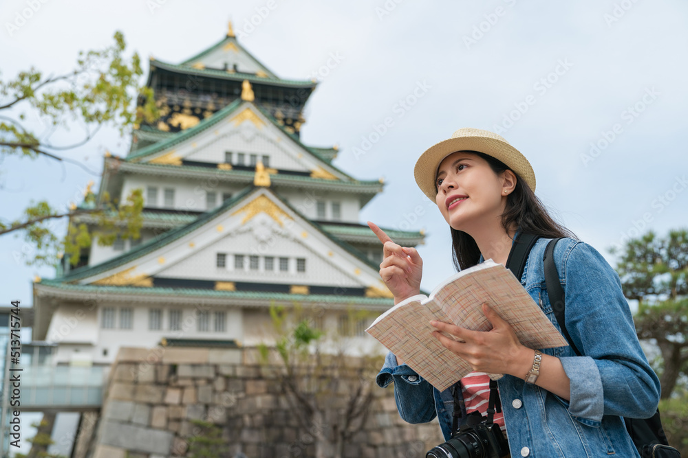 low angle shot portrait of cheerful Asian female tourist finger pointing into distance while exploring in Osaka castle park in japan with travel guidebook in hand