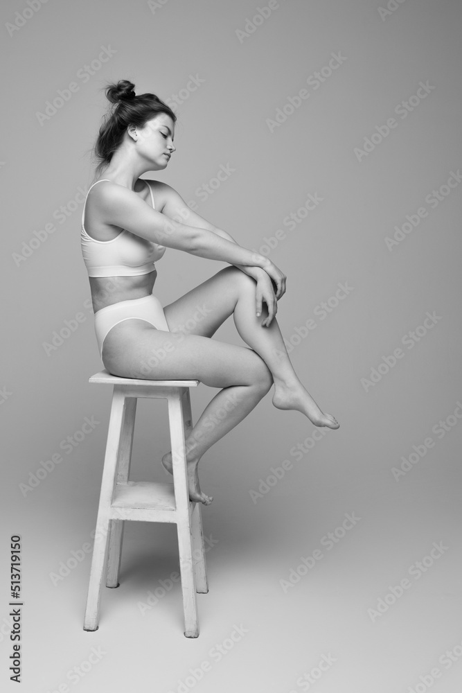 Fototapeta premium Studio portrait of young beautiful woman with slim body sitting on chair, posing in underwear. Black and white photography