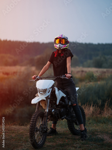 A beautiful young happy woman in a helmet poses sitting on a cross-country motorcycle at sunset photo