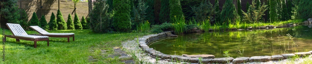 Lounge chairs on the backyard near the pond in a beautiful garden. Two empty sunbeds on the grass. Lounge chairs on the backyard near the pond in a beautiful garden. Two empty sunbeds on the grass.