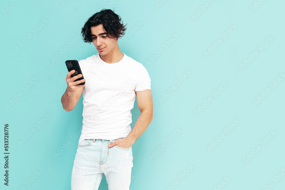 Handsome confident stylish hipster lambersexual model.Man dressed in over size T-shirt and jeans. Fashion male posing near blue wall.Holding smartphone. Looking at cellphone screen. Using apps Handsome confident stylish hipster lambersexual model.Man dressed in over size T-shirt and jeans. Fashion male posing near blue wall.Holding smartphone. Looking at cellphone screen. Using apps