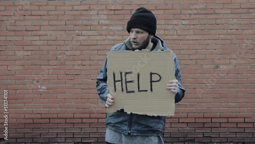 Homeless holds piece of cardboard with inscription Help. Refugee is at brick wall looking for job. Male tramp in dirty clothes, hat. Below poverty line. Bearded fugitive beggar. Immigrant. Drunkenness