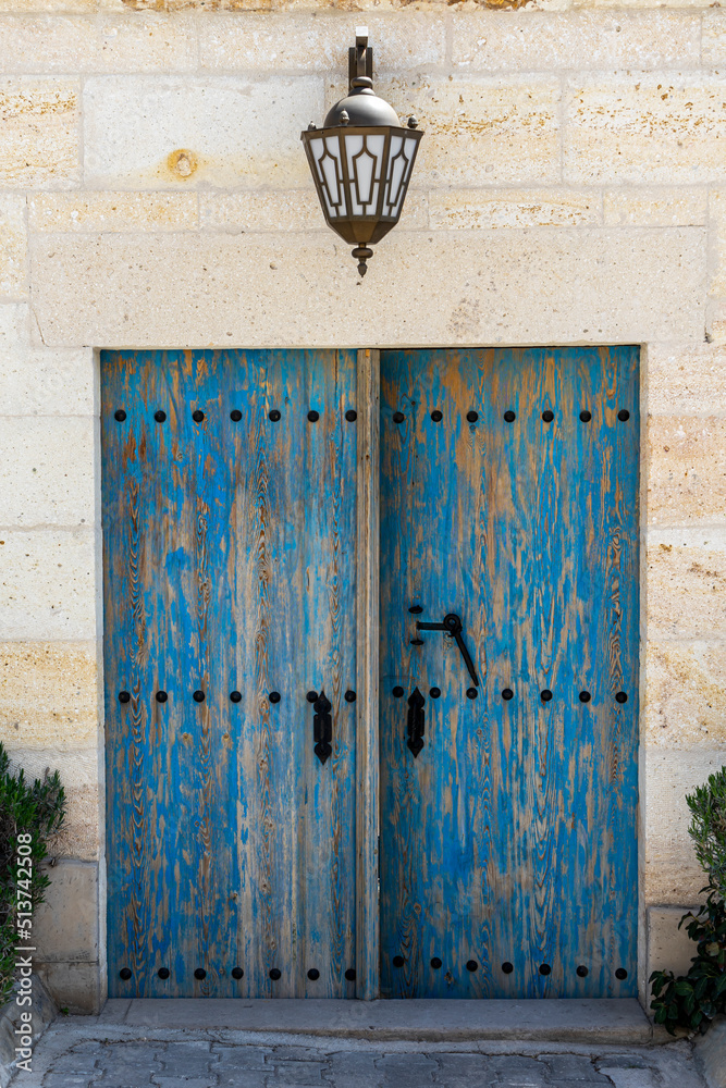 An old looking wooden blue door and decorative stone wall with a big rustic wall lamp in Ortahisar, Cappadocia, Nevsehir. An old looking wooden blue door and decorative stone wall with a big rustic wall lamp in Ortahisar, Cappadocia, Nevsehir.