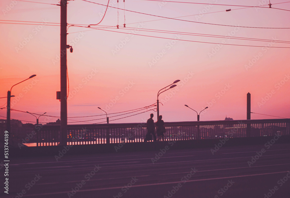 A young couple in love is walking on the bridge against the background of lanterns, wires and a pink sunset sky. The city is falling asleep. Romance. Blur