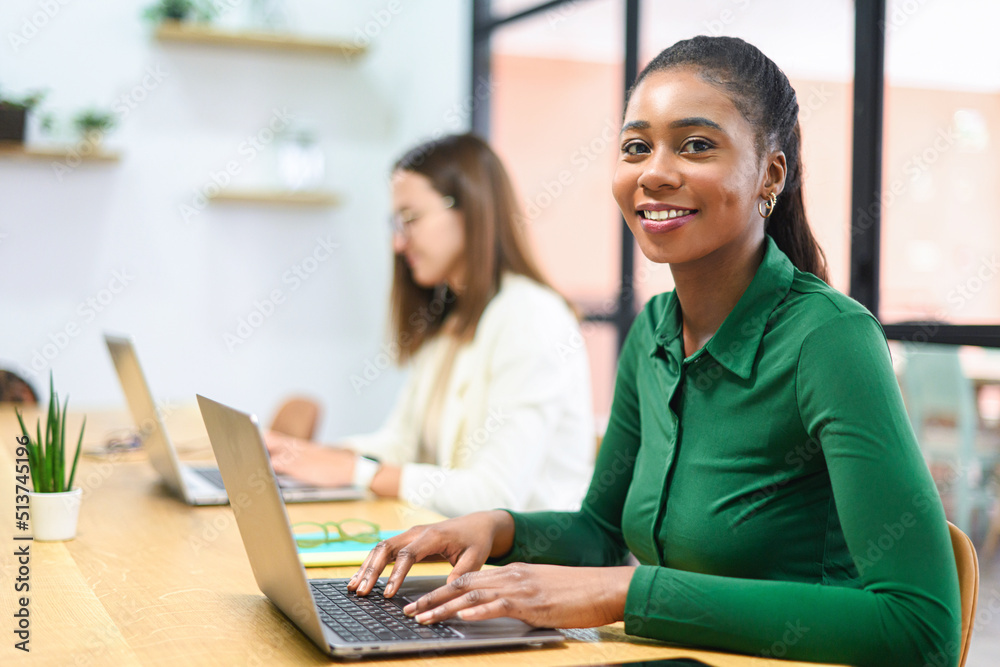 Portrait of smiling African American businesswoman with female colleague working on background, female office employees using laptops sitting in modern office space, diverse work team
