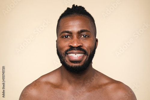 Handsome african-american young man shirtless naked looking at camera smiling with toothy smile isolated in beige background. Male beauty concept